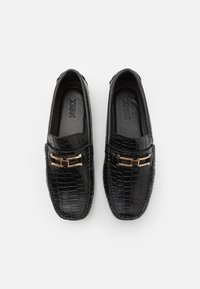 Mocassins noirs avec une texture crocodile, détails en métal doré, bout rond et semelle intérieure rembourrée pour le confort. Logo visible à l'intérieur.