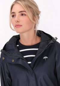 Marinblå vattentät jacka med huva, som har en silverfärgad paraplylogotyp, gyllene tryckknappar och en dragkedja framtill, bärs över en randig topp.