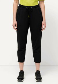 Zwarte joggerbroek met een trekkoord in de taille; voorzien van groene accenten aan de zijkanten; cropped lengte; zachte textuur; gecombineerd met zwarte sneakers.