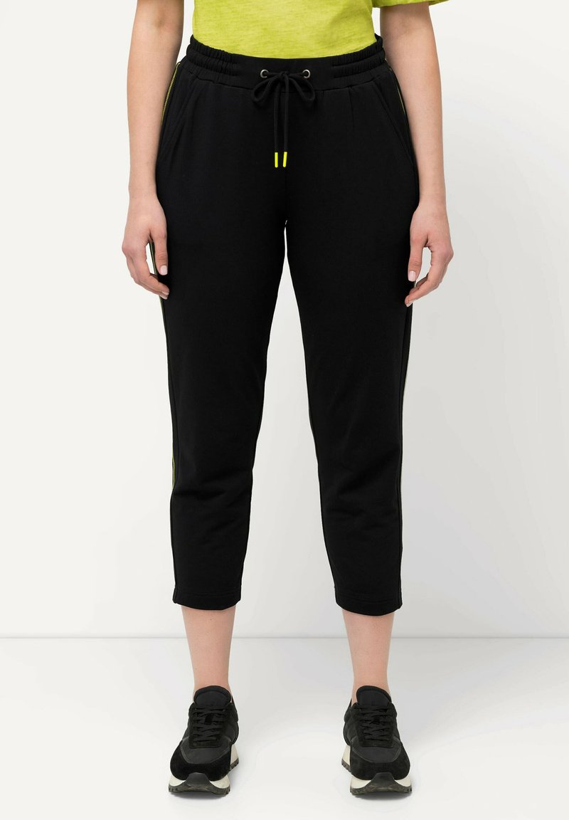 Zwarte joggerbroek met een trekkoord in de taille; voorzien van groene accenten aan de zijkanten; cropped lengte; zachte textuur; gecombineerd met zwarte sneakers.