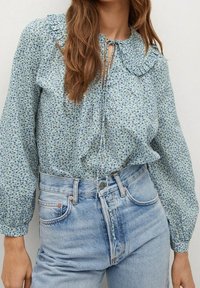 Blumenmuster-Bluse mit Knöpfen, frilltem Kragen, langen Ärmeln und einer Bindeschleife. Hat ein blaues und grünes Muster auf einem hellen Hintergrund.