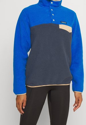 Personne portant un pull polaire bicolore bleu et gris avec bordure beige, boutons-pression et petite poche poitrine à gauche.