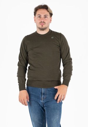 Maglione lavorato a maglia verde oliva con maniche lunghe, colletto girocollo e polsini a costine. Presenta un piccolo logo sul lato sinistro del petto. Indossato con jeans blu.
