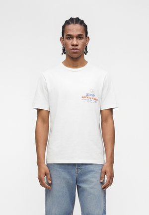 Noormees palmikutega, kes kannab valget Jack & Jones t-särki ja hele siniseid teksapükse, seisab valge ühevärvilise tausta ees.