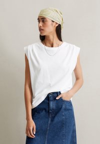 Witte katoenen mouwloze top met een relaxte pasvorm en ronde halslijn, gecombineerd met een blauwe denim rok. Het model draagt een lichtgroene geprinte hoofddoek.