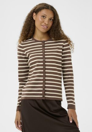 Vrouw met krullend haar die een bruine en beige gestreepte cardigan met knopen en een donkerbruine rok draagt, poserend tegen een effen achtergrond.