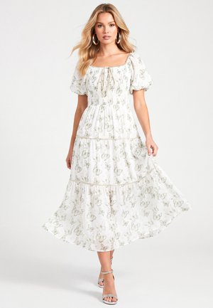 Femme en robe blanche longue jusqu'aux chevilles avec des manches bouffantes et un imprimé papillon, portant des sandales argentées à talons hauts, debout sur un fond uni.