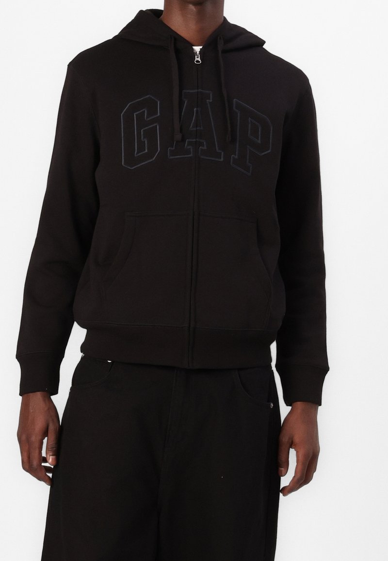 Personne portant un sweat à capuche noir zippé avec capuche et grandes poches frontales, avec un logo GAP embossé sur la poitrine.