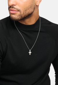 Silberne Kreuzkette an einer dünnen Kette, getragen über einem schwarzen, gerippten Langarmshirt. Das Kreuz zeigt ein einfaches, modernes Design.