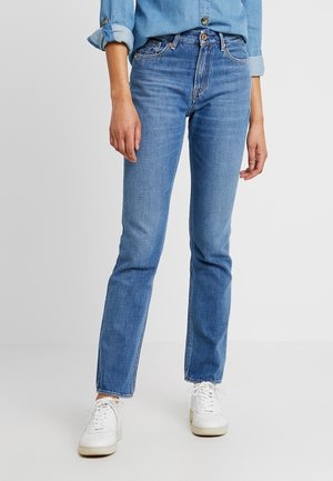 Person trägt hellblaue Jeans mit geradem Bein, hochgekrempelte Ärmel eines Jeanshemds und weiße Turnschuhe, steht vor einem einfarbigen Hintergrund.