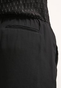 Pantalones negros de tela suave con cintura elástica, que cuentan con un bolsillo lateral discreto y un sutil detalle texturizado por encima de la cintura.
