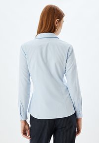 Camicia lunga azzurro chiaro con colletto classico, design aderente e polsini abbottonati. Tessuto morbido con una leggera lucentezza.