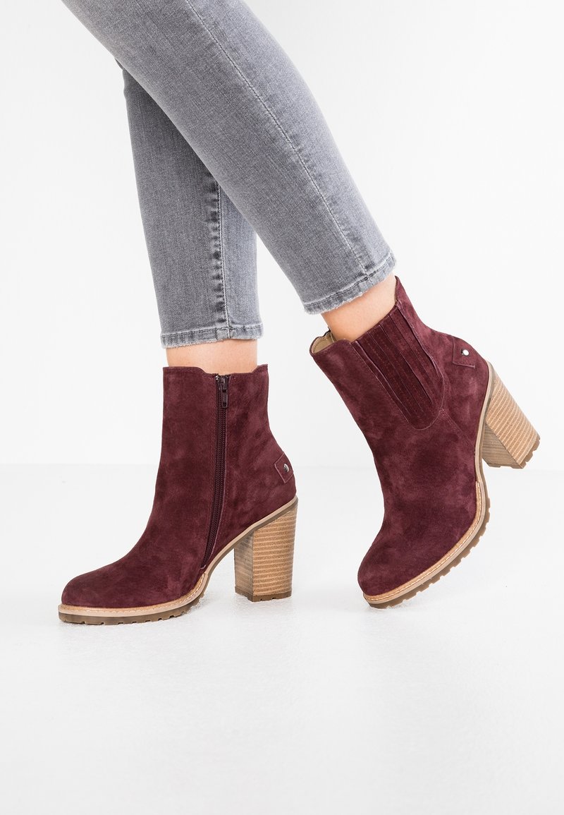 Personne portant un jean gris ajusté et des bottines bordeaux en daim avec talons blocs en bois empilés et fermeture éclair latérale.