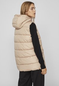 Gilet rembourré beige avec capuche, texture matelassée, poches latérales zippées et coupe ample. Porté sur un haut noir à manches longues.