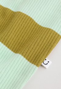 Tela acanalada con franjas horizontales anchas en verde claro y amarillo mostaza, con una pequeña etiqueta blanca con una cara sonriente negra.
