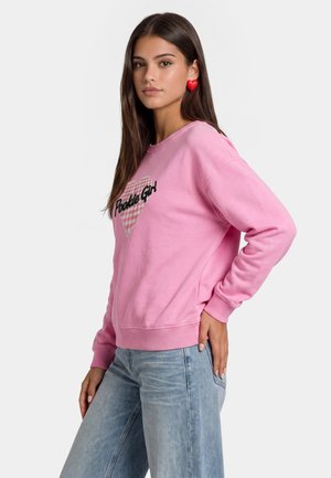 Młoda kobieta ubrana w różową bluzę z napisem "Pookie Girl" i grafiką serca, niebieskie dżinsy oraz czerwone kolczyki w kształcie serca.