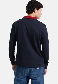 Maglietta a maniche lunghe navy con costine, colletto rosso, scollo rotondo e vestibilità rilassata, indossata con jeans denim blu, vista da dietro.