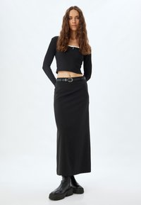 Zwarte geribbelde lange mouwen crop top gecombineerd met een zwarte midi rok. De rok heeft een gladde textuur en een riem met een rechthoekige gesp. Zwarte laarzen.