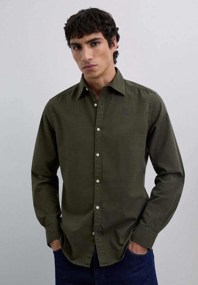 Young man wearing olive green checkered button-up shirt and dark jeans, standing with hands in pockets against plain background.