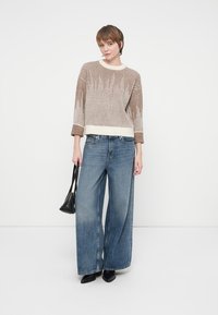 Beige og creme mønstret striktrøje med ribbet tekstur, parret med brede blå jeans. Modellen holder en lille sort taske og har sorte sko på.
