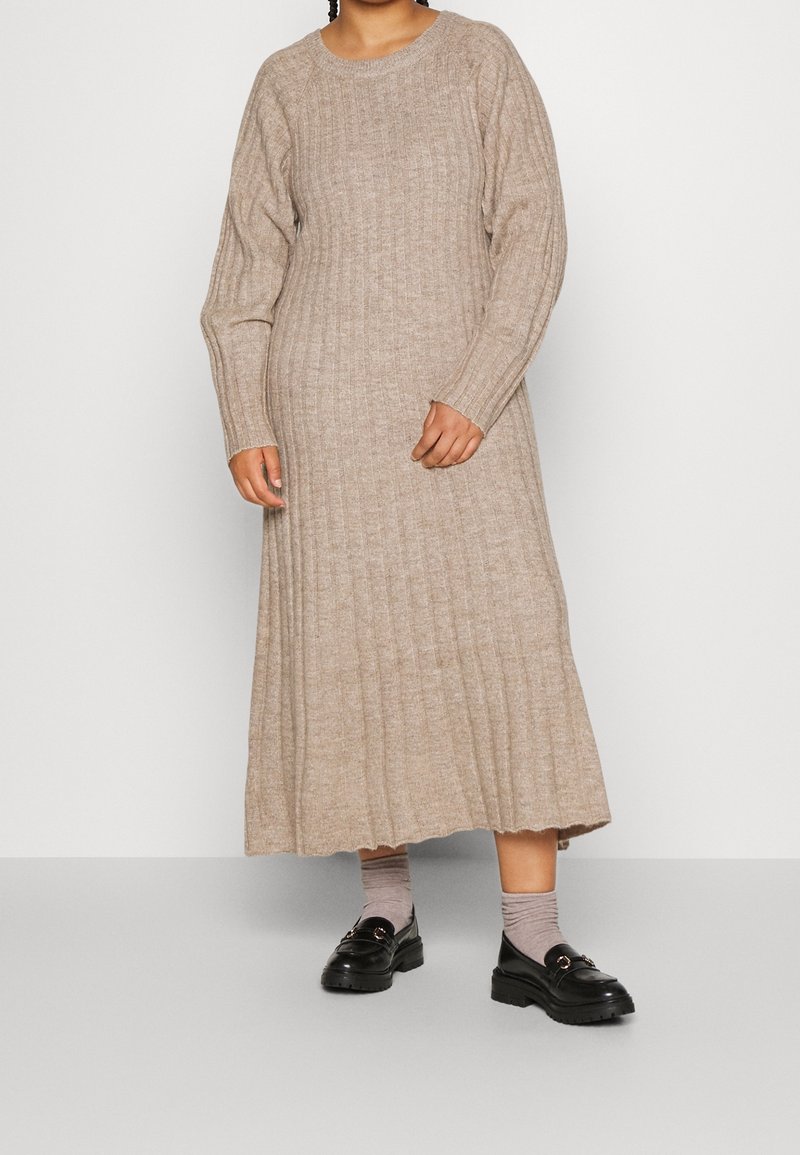 Femme portant une longue robe en tricot côtelé beige à manches longues, associée à des chaussettes grises et des mocassins noirs.