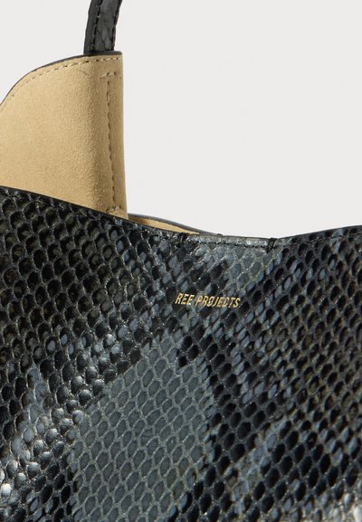 Primo piano di una scarpa nera in pelle texturizzata con dettagli in suede beige e il logo dorato "REE PROJECTS" impresso di lato.