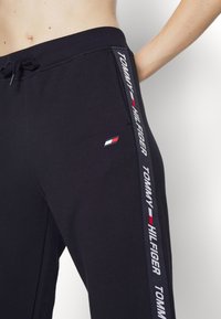 Schwarze Jogginghose aus weichem Material, mit einem Kordelzugbund, seitlichen Logo-Elementen und einem weißen Textstreifen entlang des Beins.