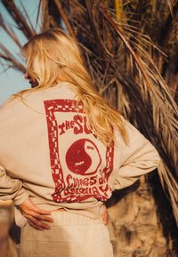 Beżowa bluza z nadrukiem w czerwonym kolorze przedstawiającym słońce oraz tekstem "słońce wschodzi" z bordiurą w szachownicę. Miękki materiał, luźny krój.