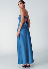 Mėlyna satino ilga suknelė su nugaros išpjova ir surinkta petneša. Lygus paviršius, grindis siekianti ilgio, dėvima su juodais aukštakulniais bateliais.