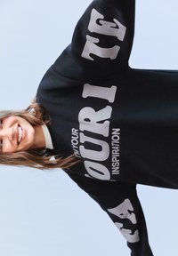 Čierna mikina s veľkým sivým textom "TVOJ INŠPIRÁCIA" a "OBĽÚBENÉ" v tučnom písmene. Má uvoľnený strih a rebrovaný golier.