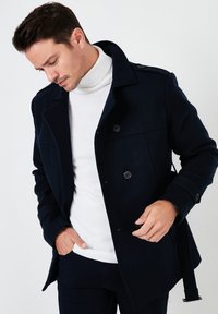 Sötétkék gyapjú keverék kabát, kifli gallérral, gombos záródással és oldalsó zsebekkel; fehér garbó pulóverre viselve, amelynek letisztult a dizájnja.