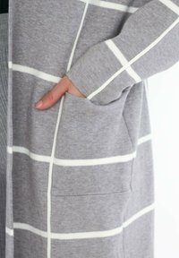 Cardigan gris avec des rayures horizontales blanches, texture en tricot et une poche avant sur le côté gauche, avec des manches à coupe relaxée.