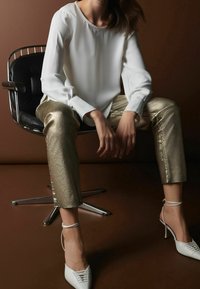 Weiße Bluse mit langen Ärmeln, metallisch goldene, kurze Hose und weiße High Heels mit Spitzendetails. Sitzt auf einem schwarzen Stuhl vor einem braunen Hintergrund.