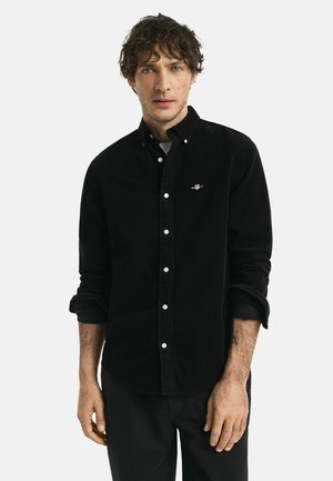 Zwarte corduroy shirt met een button-down kraag, witte knopen en een klein logo op de borst, gecombineerd met zwarte broek. Zachte textuur.