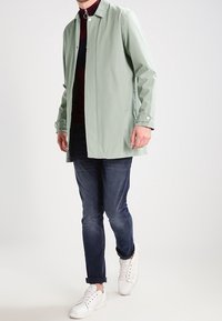 Mann trägt eine mintgrüne Jacke, dunkle Jeans, weiße Sneaker und ein bordeauxrotes Oberteil, steht vor einem schlichten hellen Hintergrund.