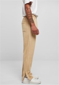 Beige sweatpants lavet af blødt stof, med en sidelomme, smal pasform og detaljer ved kanten, parret med hvide sneakers.