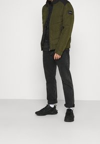 Olivengrøn puffjakke med sorte detaljer, lynlåslukning og ståkrave, kombineret med sorte jeans og sorte sneakers. Hvide sokker er synlige.