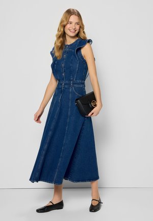 Denim jurk met een aansluitende lijfje, gerimpelde mouwen en een lange rok. Donkerblauwe stof, knopen-voorpand detail en ceintuur taille. Zwarte handtas.