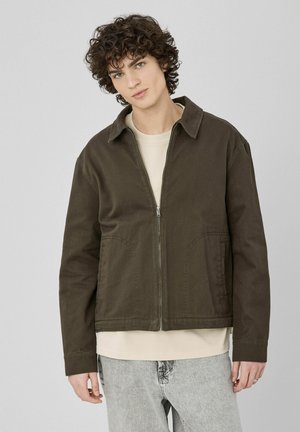 Jeune homme aux cheveux bouclés portant une veste zippée marron foncé, une chemise beige et un jean gris clair sur un fond clair uni.