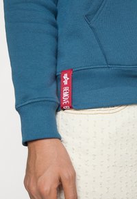 Felpa blu con polsini a costine presenta un'etichetta rossa con marchio sull'orlo. Il materiale sembra morbido con una texture liscia.