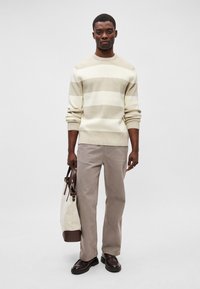 Gestreifter Pullover in Creme und Beige, graue Hose, dunkle Schuhe, hält eine zweifarbige Tasche mit sandfarbenen und weißen Akzenten vor einem neutralen Hintergrund.