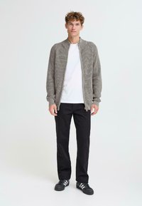 Jasnoszary sweter z dzianiny z wysokim kołnierzem, pełnym zamkiem, o ribbanym wzorze, noszony na białej koszulce i czarnych spodniach, w zestawieniu z czarnymi trampkami.