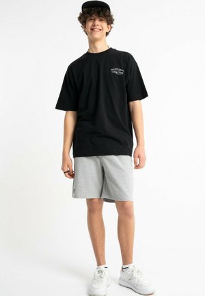 Uśmiechający się młody mężczyzna w czarnej czapce, czarnej oversize koszulce, szarych szortach i białych trampkach na jednolitym białym tle.