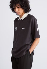 Polo negro con un cuello beige en contraste, que presenta un emblema en el lado izquierdo y un número "1" blanco en la manga. Textura suave.