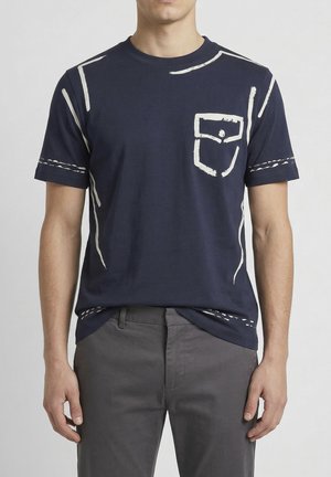 Hombre con camiseta azul marino con diseños de líneas blancas que simulan un bolsillo y costuras, combinada con pantalones grises.