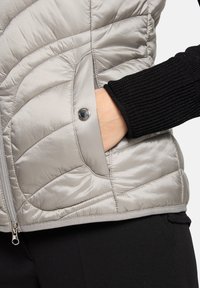 Veste matelassée gris clair avec une texture lisse et brillante. Comprend des manches côtelées noires et un détail de bouton-pression sur la poche.