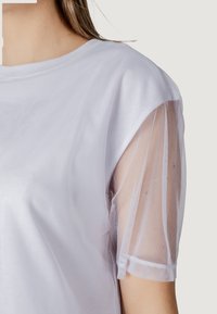 Top blanco con mangas de malla transparente, escote redondo y textura suave. Los detalles destacables incluyen un tejido ligero y acentos transparentes en las mangas.