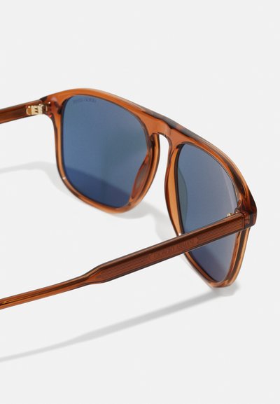 Lunettes de soleil brunes transparentes avec des verres bleus, des montures épaisses et des détails gravés sur les branches. Surface réfléchissante avec une texture lisse.