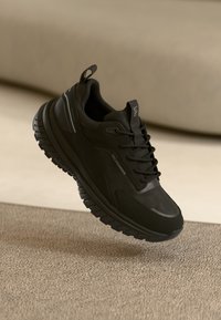 Sapatilha esportiva preta com um cabedal texturizado e leve, entressola acolchoada e sola robusta. Apresenta uma marcação discreta e detalhes refletivos.