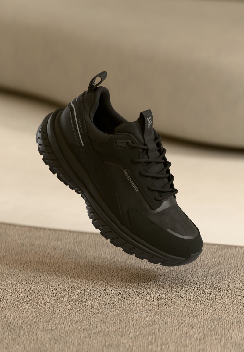 Sapatilha esportiva preta com um cabedal texturizado e leve, entressola acolchoada e sola robusta. Apresenta uma marcação discreta e detalhes refletivos.